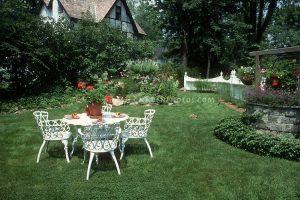 Summer Garden with white wrought iron furniture table and chairs, lawn grass and house, wishing well in yard, flowers, picket fence for beautiful backyard living